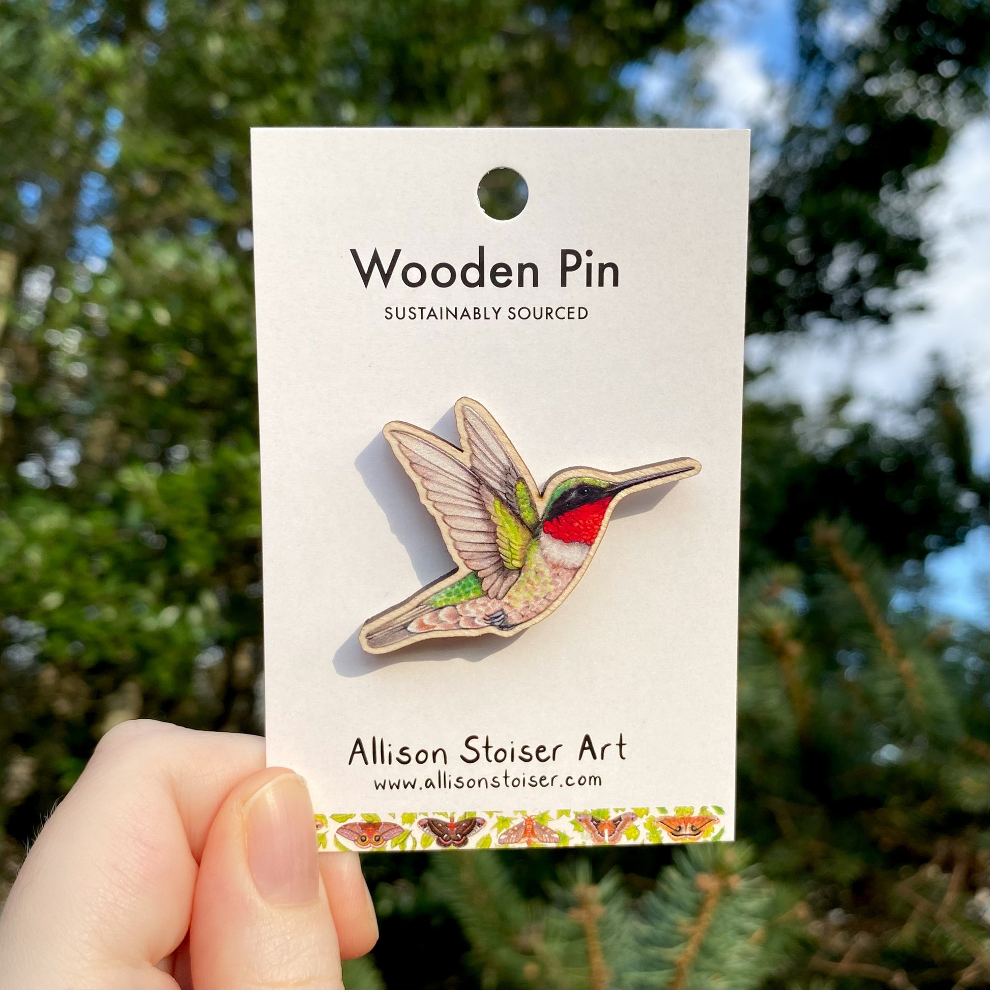 A hand holding a wooden pin with an illustration of a male ruby-throated hummingbird, sustainably sourced, with the brand name Allison Stoiser Art visible.