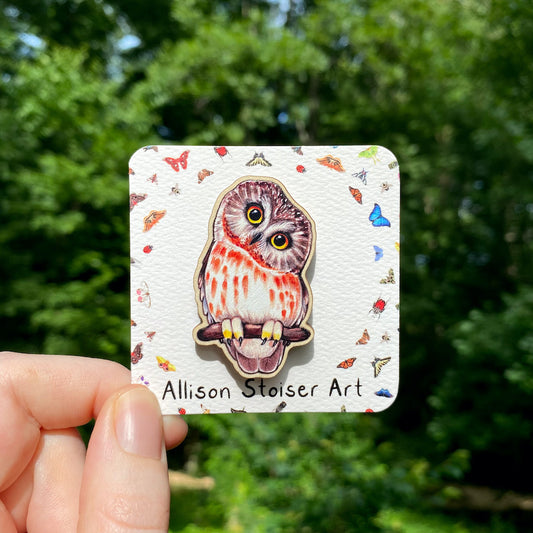 A hand holds a sustainably sourced wooden pin featuring an illustration of a saw-whet owl