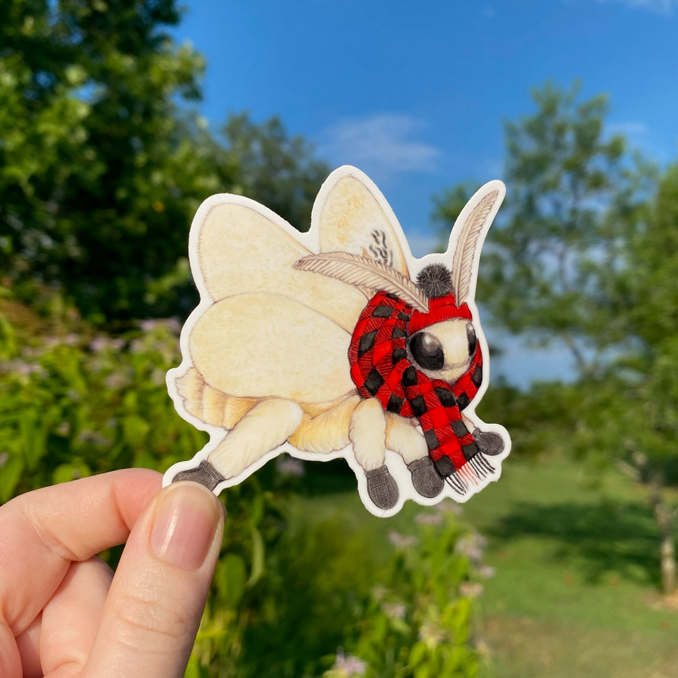 A hand holding a sticker of a flannel moth wearing a red flannel scarf.