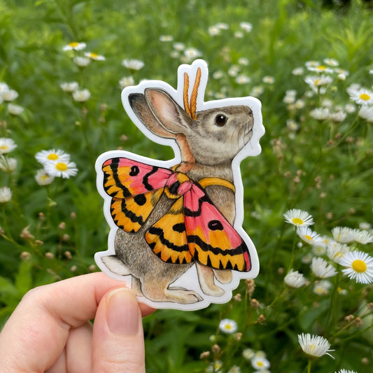 Hand holding a sticker of a rabbit with sheep moth wings against a natural background with flowers.