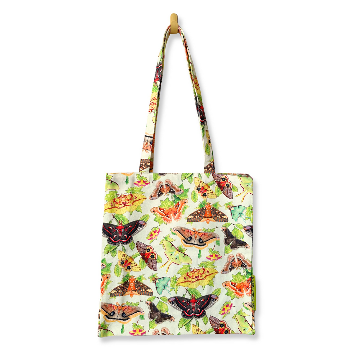 A canvas tote bag with a white background covered in a pattern of moths and leaves, hanging from a wooden peg on a black background.