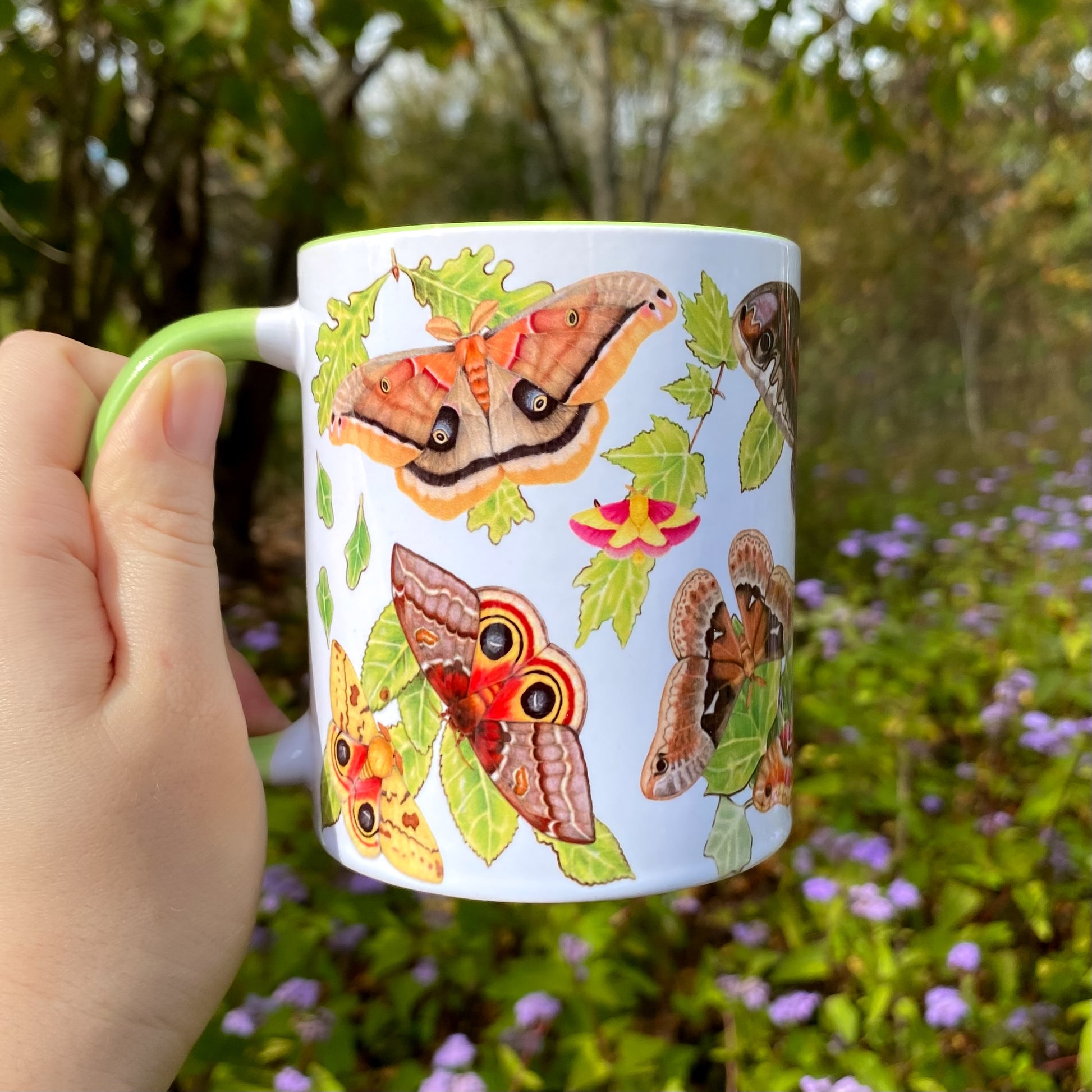 Mug with silk moth illustrations held by a hand in a natural setting with flowers and trees.
