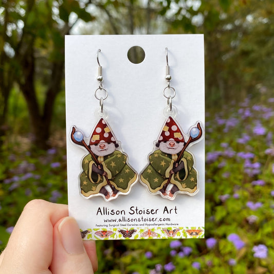 Pair of wizard opossum earrings on a card held in front of a natural background with flowers.