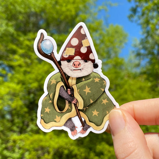 A hand holding a vinyl sticker with an illustration of a cute opossum dressed as a sorcerer with a hat, staff, and a green robe with gold stars.