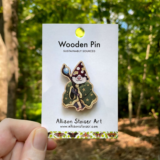 Wooden pin of a wizard opossum on a card with 'Allison Stoiser Art' branding, held against a forest background.