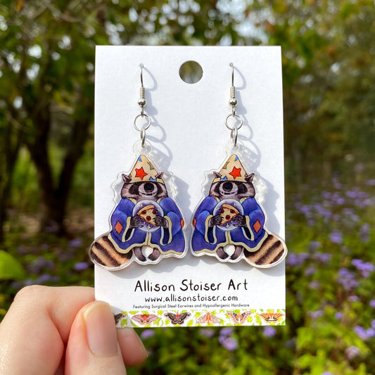 Raccoon wizard earrings on a card held in front of a natural background