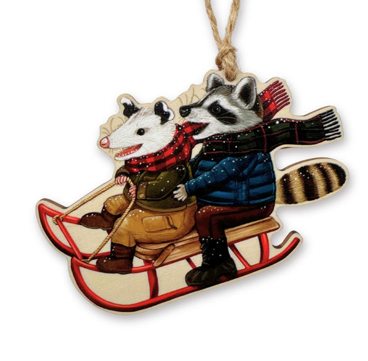 Decorative ornament with an opossum and raccoon on a sled, wearing scarves and coats.