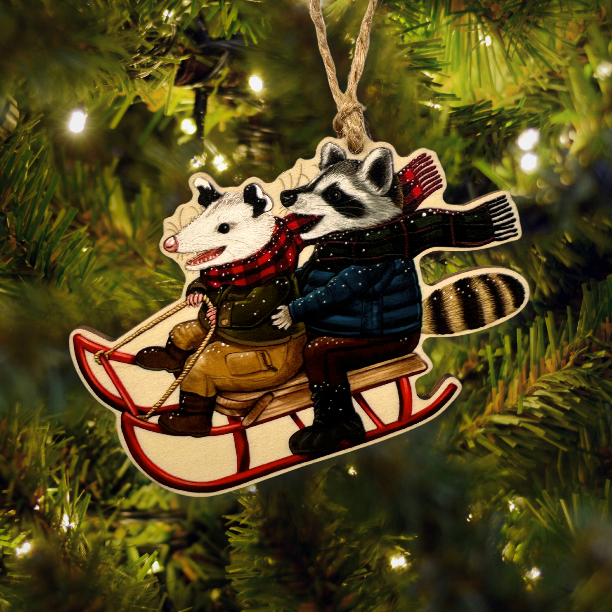 Decorative ornament with a raccoon and opossum on a sled hanging on a Christmas tree.