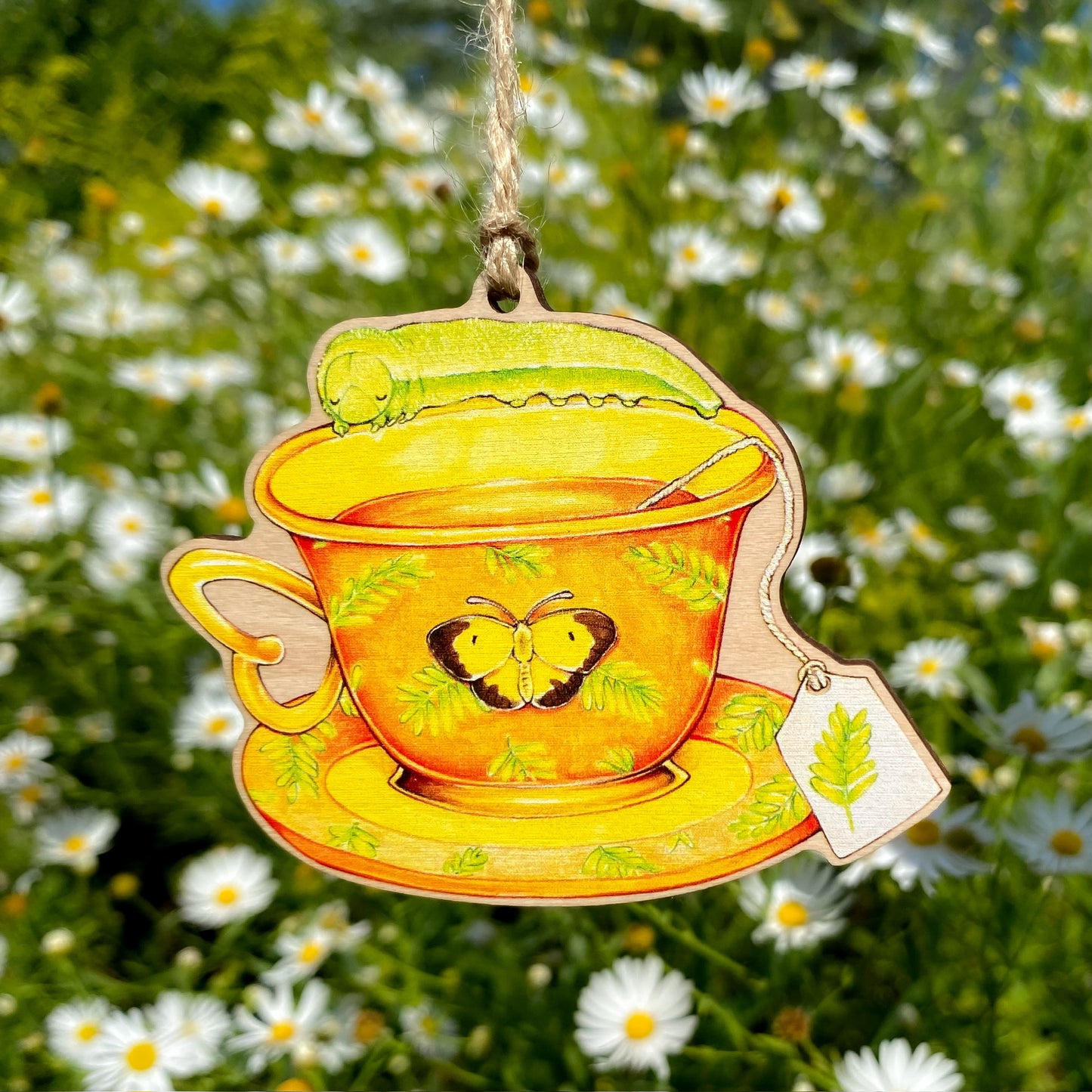 Decorative wooden ornament of a teacup with a green sleepy orange caterpillar hanging against a natural background with flowers.