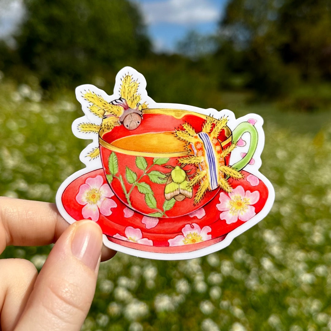 A handholding a sticker of a red teacup with rose motifs with spiky yellow and red caterpillars drinking from it.