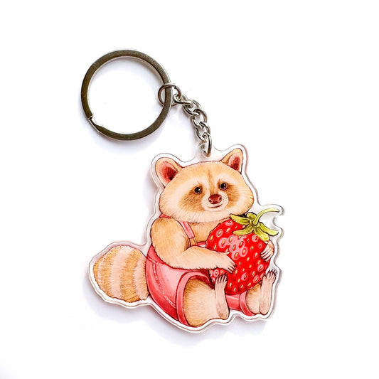 A keychain of a blond raccoon in pink overalls holding a strawberry on a white background.