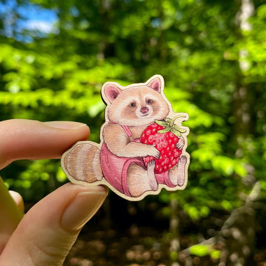 A hand holding a wooden pin of a blond raccoon in pink overalls holding a big strawberry.