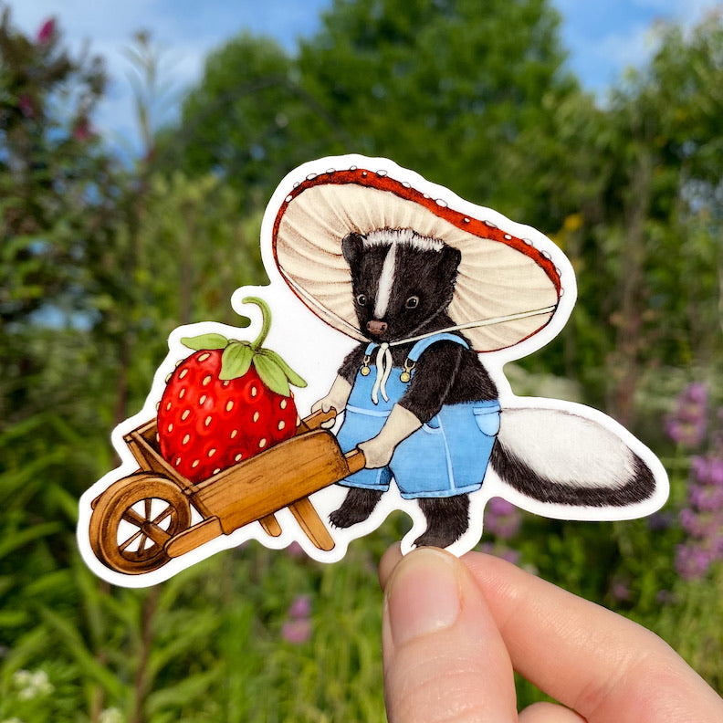 A hand holding a sticker of a skunk in overalls pushing a wheelbarrow with a big strawberry.