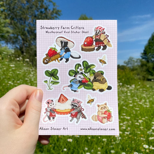 A hand holding a pink sticker sheet that reads "Strawberry Farm Critters Weatherproof Vinyl Sticker Sheet" along the top and "Allison Stoiser Art" and "www.allisonstoiser.com" along the bottom. The stickers include a badger cutting a strawberry, a skunk with a wheelbarrow and strawberry, a strawberry plant, a baby opossum and beaver picking a strawberry, bees, an opossum and raccoon holding a cake with a strawberry on top, and a raccoon in pink overalls holding a strawberry.