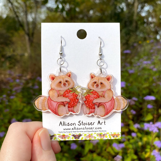 Earrings of raccoons holding strawberries on a card with 'Allison Stoiser Art' branding, held against a natural background.