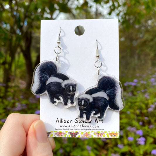 Skunk earrings on a card held in front of a natural background with greenery and flowers.