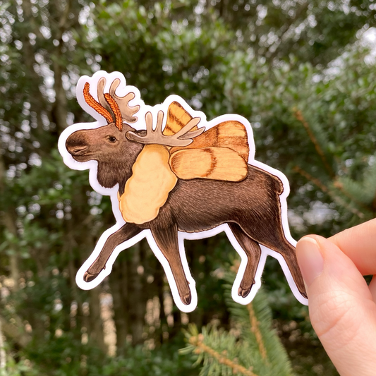A hand holding a die-cut gloss vinyl sticker of a moose with tent moth wings, against a blurred natural background.
