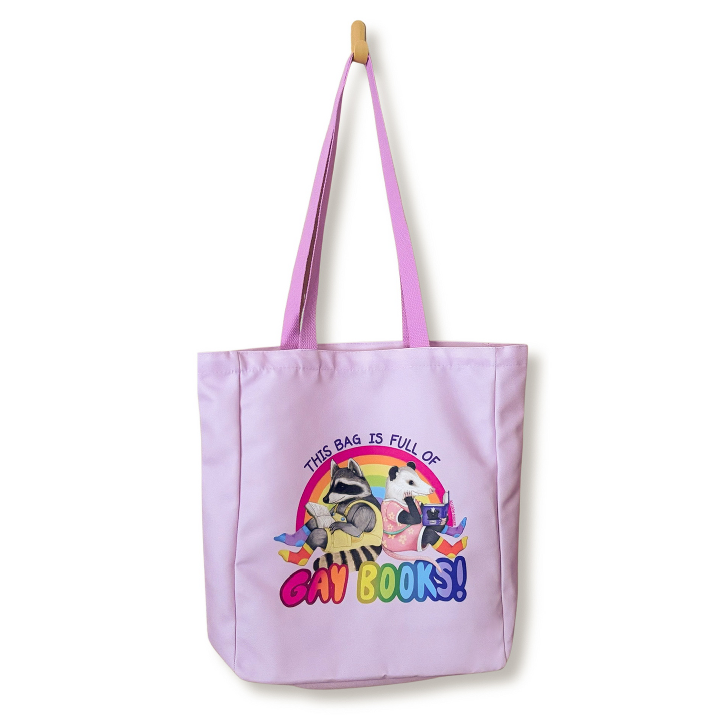 A light purple canvas tote bag with pink handles, featuring a raccoon and opossum over the text 'THIS BAG IS FULL OF GAY BOOKS!' on a white background.