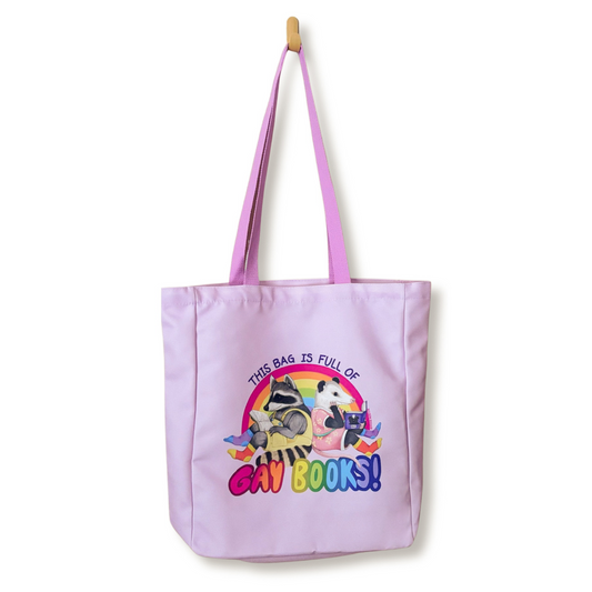 A light purple canvas tote bag with pink handles, featuring a raccoon and opossum over the text 'THIS BAG IS FULL OF GAY BOOKS!' on a white background.