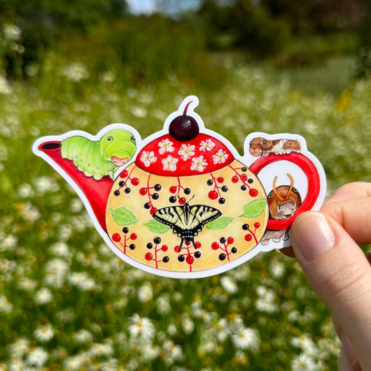 A hand holding a sticker of a yellow and red teapot surrounded by tiger swallowtail caterpillars.