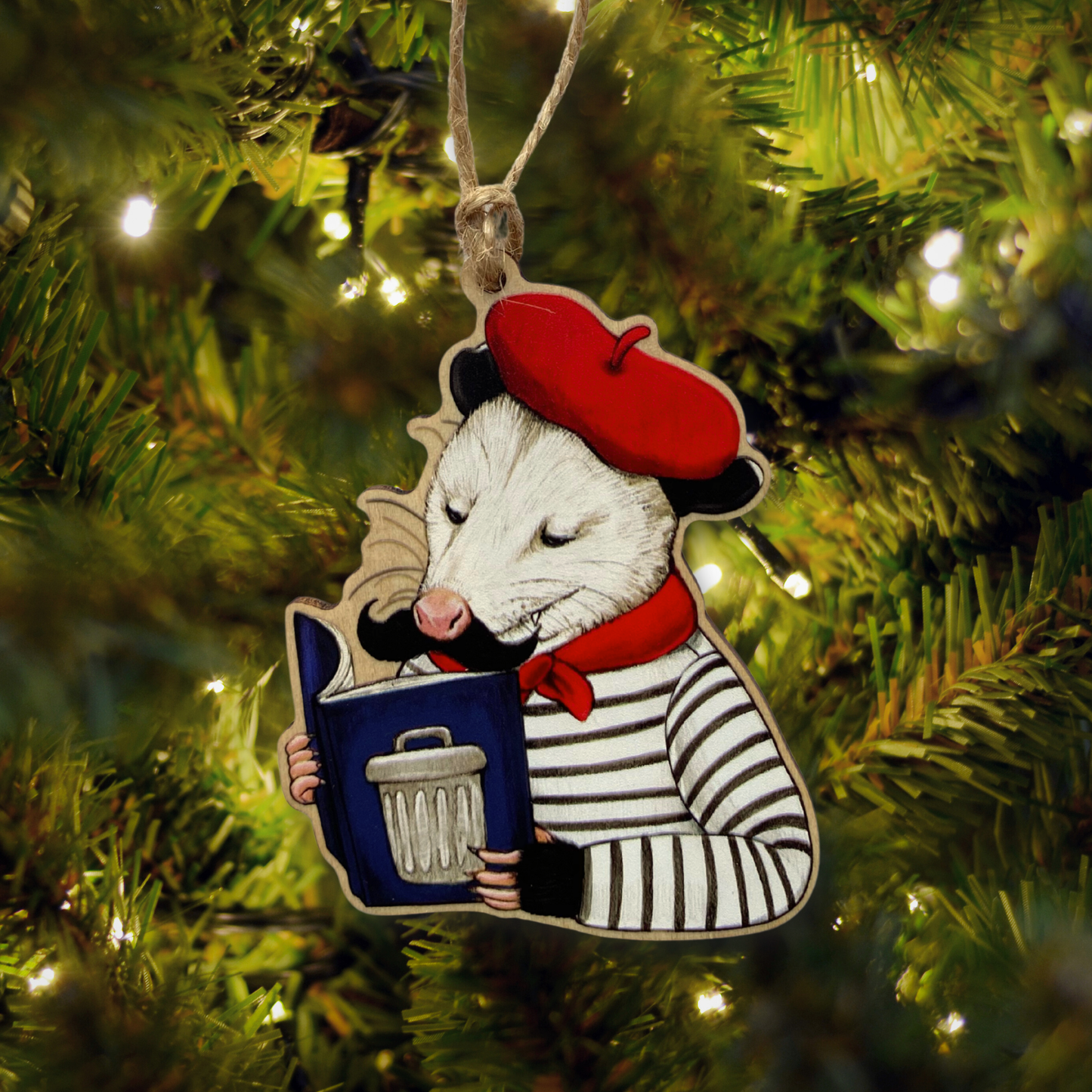 Decorative Christmas ornament of an opossum wearing a red beret and striped shirt, holding a book with a trash can on the cover, hanging on a Christmas tree.