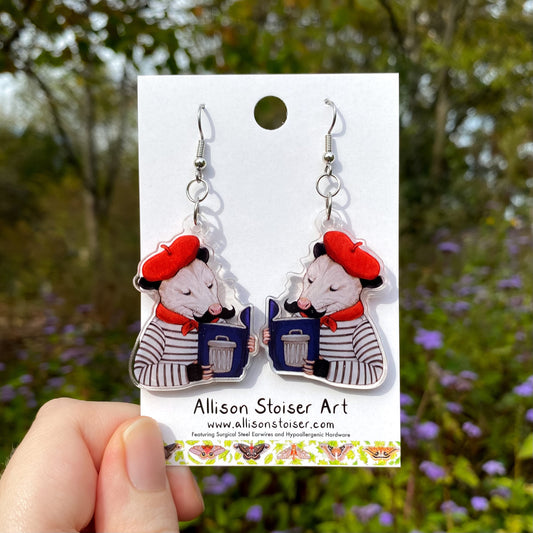 Pair of earrings featuring opossums reading books with trash cans on the cover on a card with 'Allison Stoiser Art' branding, held against a natural background.