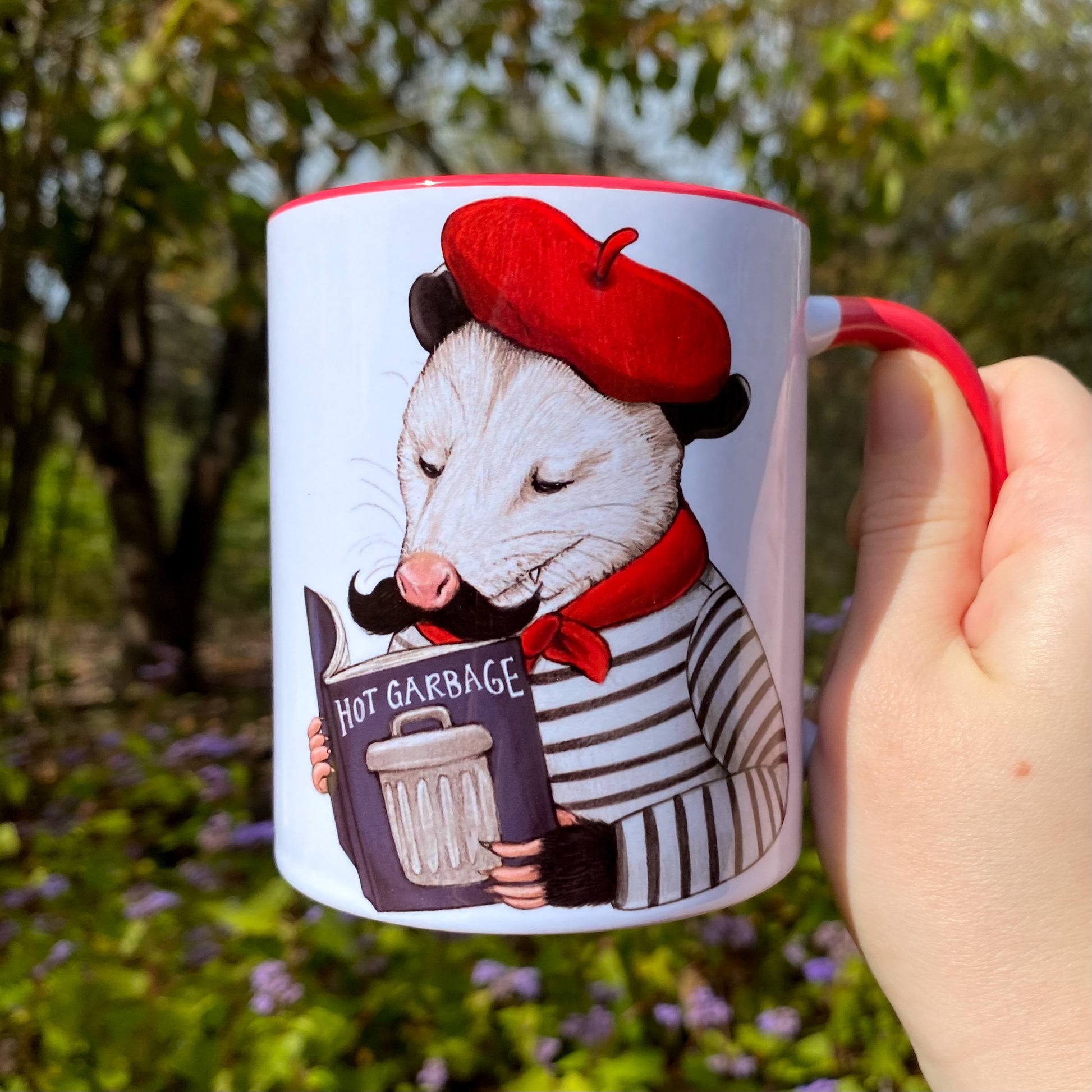 Mug with an opossum holding a book that says 'Hot Garbage' against a natural background