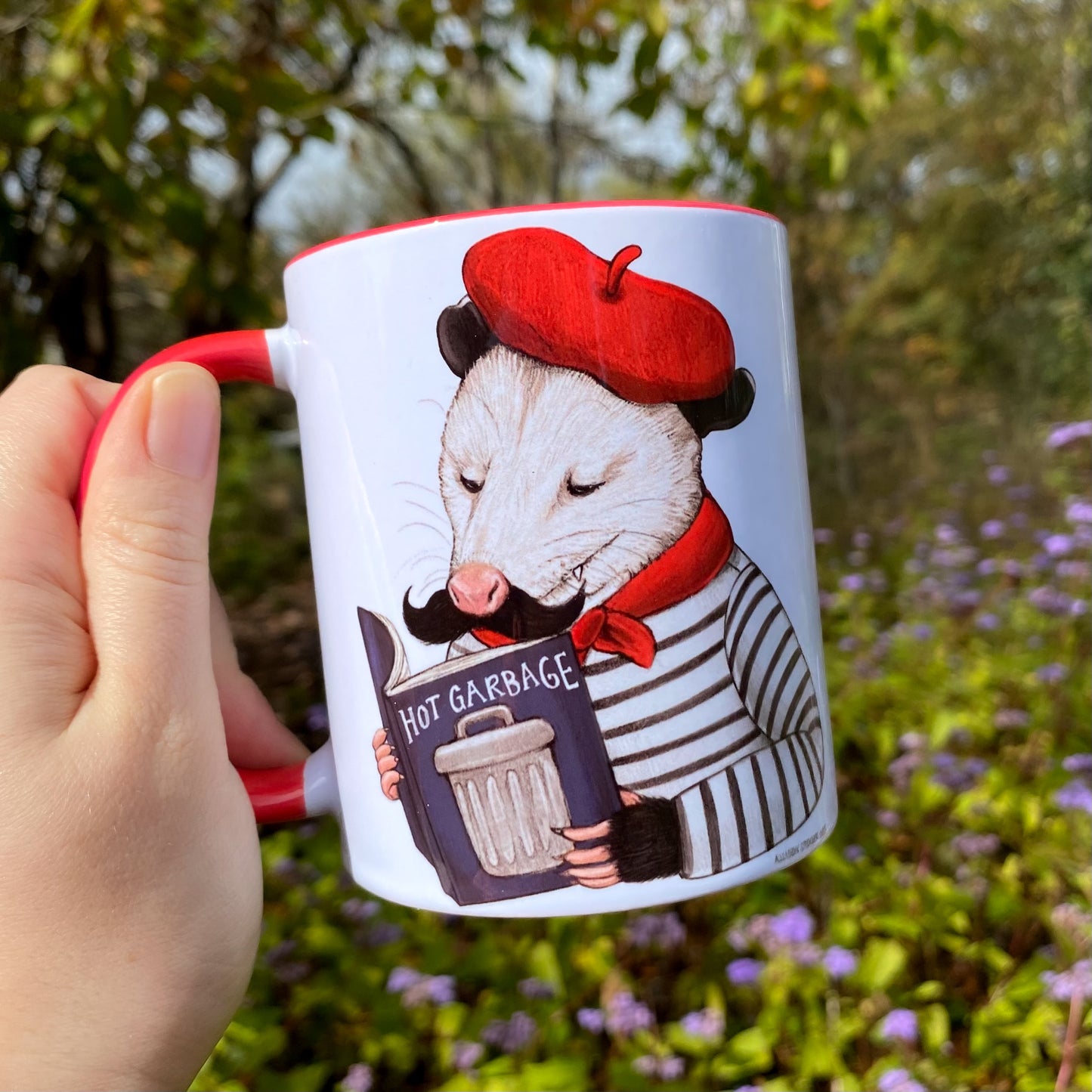 Mug with an opossum wearing a red beret and holding a book that says 'Hot Garbage' against a natural background.