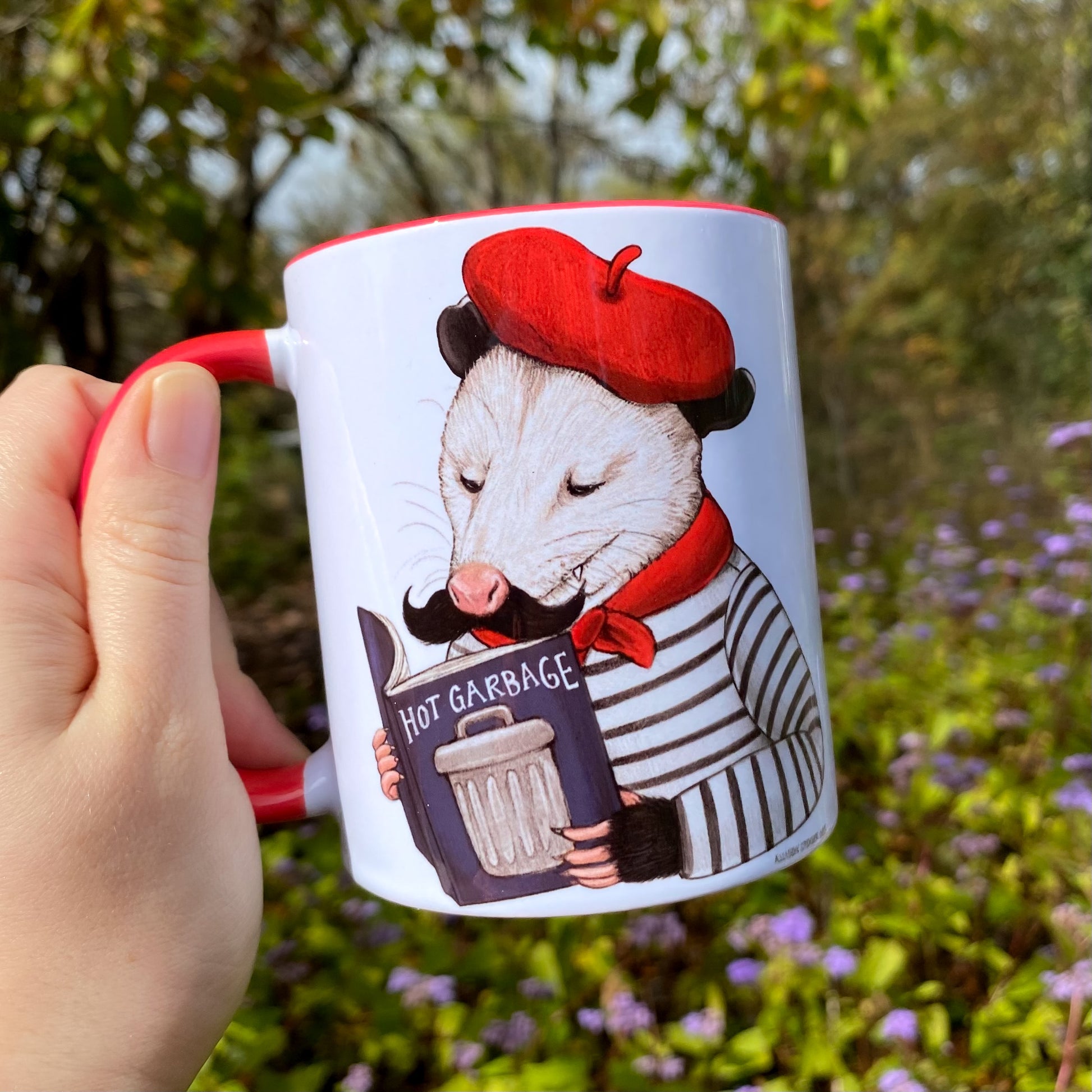 Mug with an opossum wearing a red beret and holding a book that says 'Hot Garbage' against a natural background.