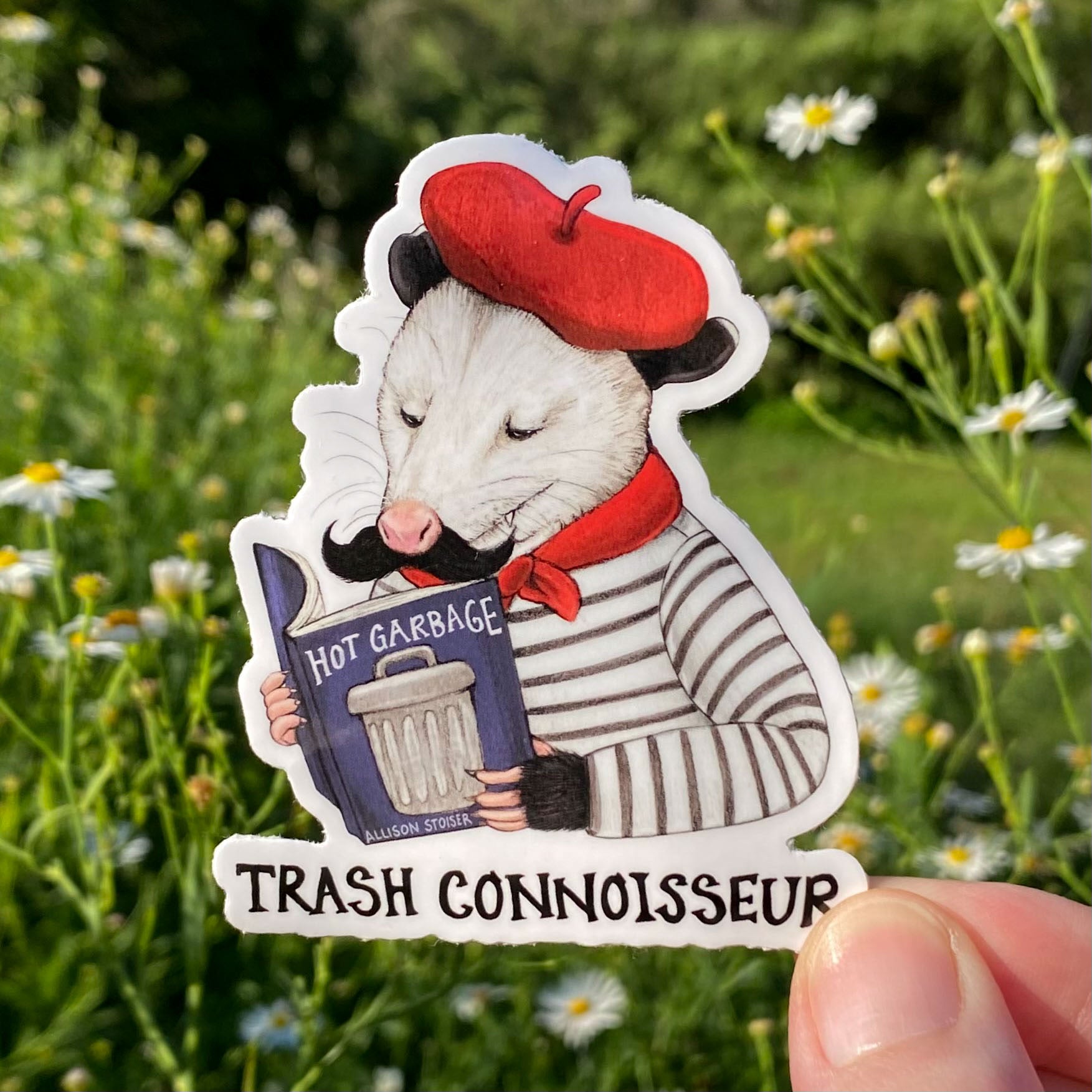 Sticker of an opossum in a red beret holding a book titled 'Hot Garbage' with text 'Trash Connoisseur' against a natural background.