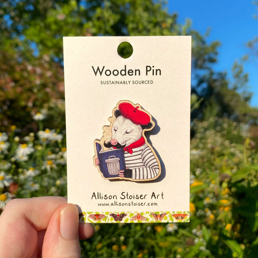 Wooden pin of an opossum reading a book titled "Hot Garbage" on a card with 'Allison Stoiser Art' branding, held against a natural background.