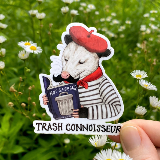 Sticker of an opossum in a beret holding a book titled 'Hot Garbage' with text 'Trash Connoisseur' against a natural background.