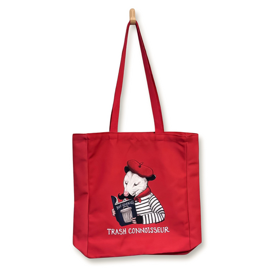 A red canvas tote bag with long shoulder straps, featuring an illustration of an opossum in a red beret and striped shirt reading a book titled Hot Garbage, with the text 'TRASH CONNOISSEUR' printed below.