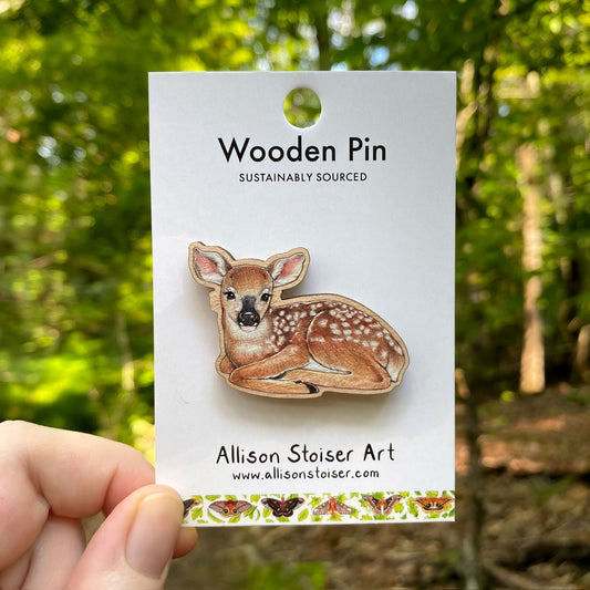 Wooden pin of a deer fawn on a card with 'Allison Stoiser Art' branding, held against a forest background.