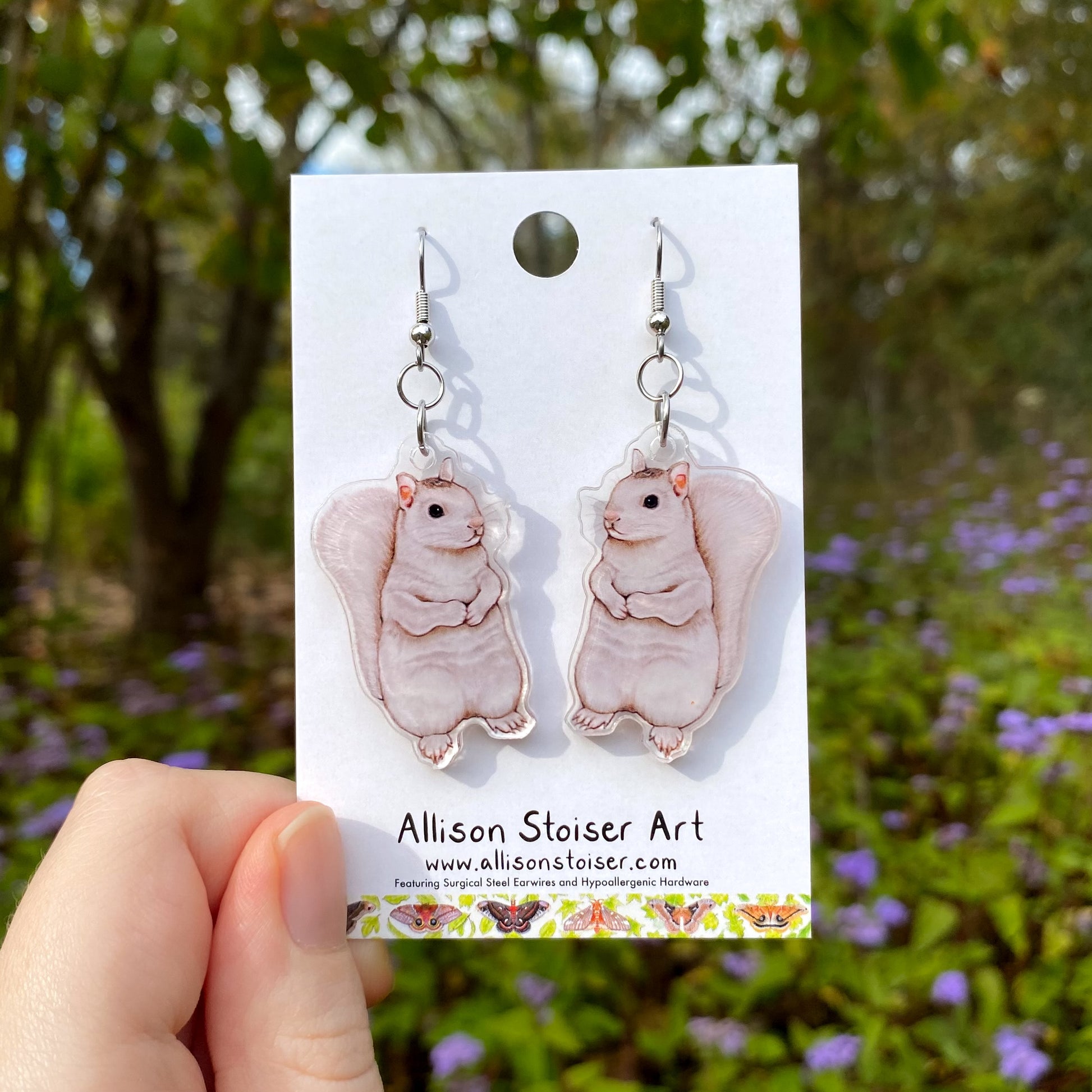 White squirrel earrings on a card held in front of a natural background with trees and flowers.
