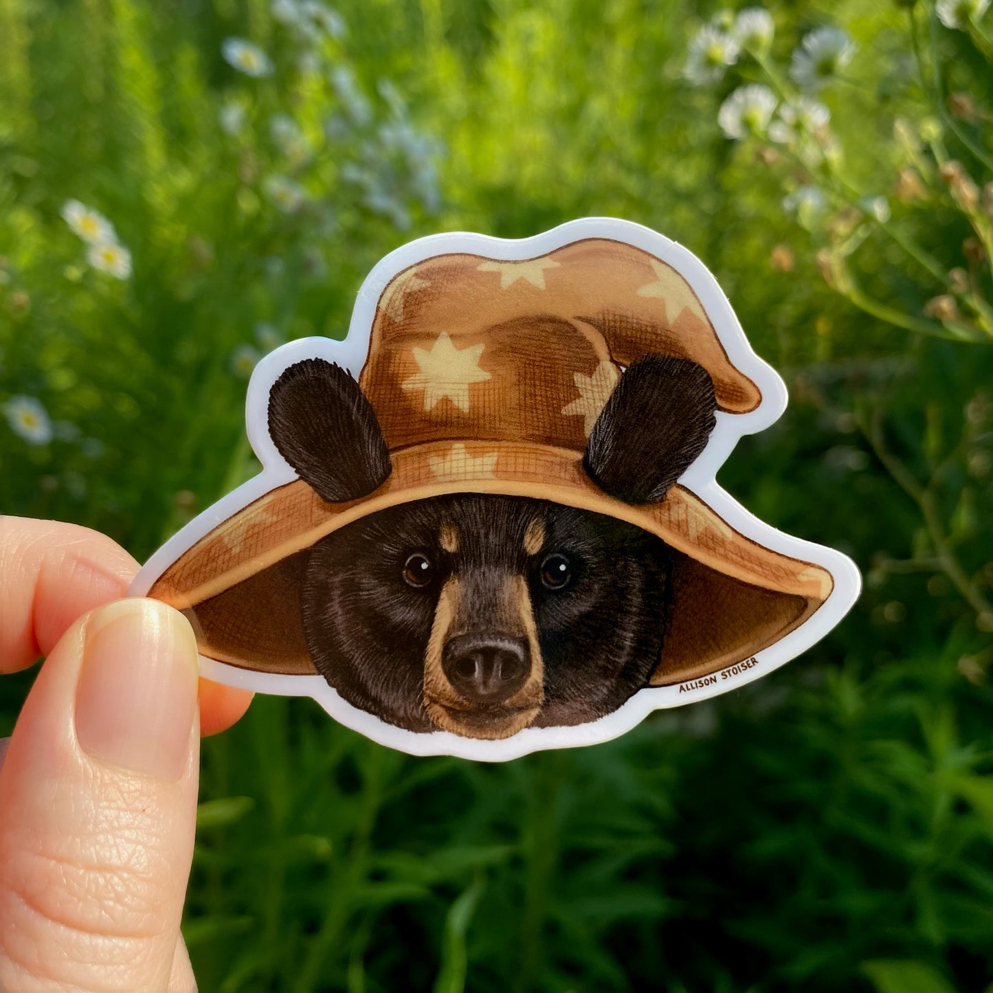 Vinyl sticker of a bear wizard with a brown hat held in front of a background with flowers