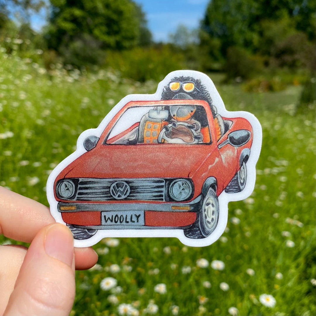 A hand holding a metallic sticker of a woolly bear caterpillar driving a red convertible.