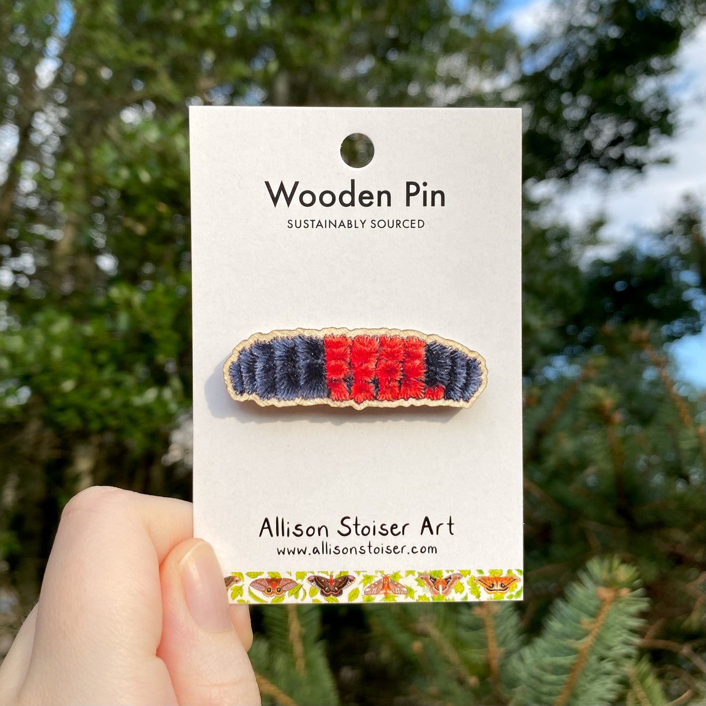 A wooden pin with a woolly bear caterpillar design, held by a hand, with a card that reads 'Wooden Pin Sustainably Sourced' from Allison Stoiser Art.