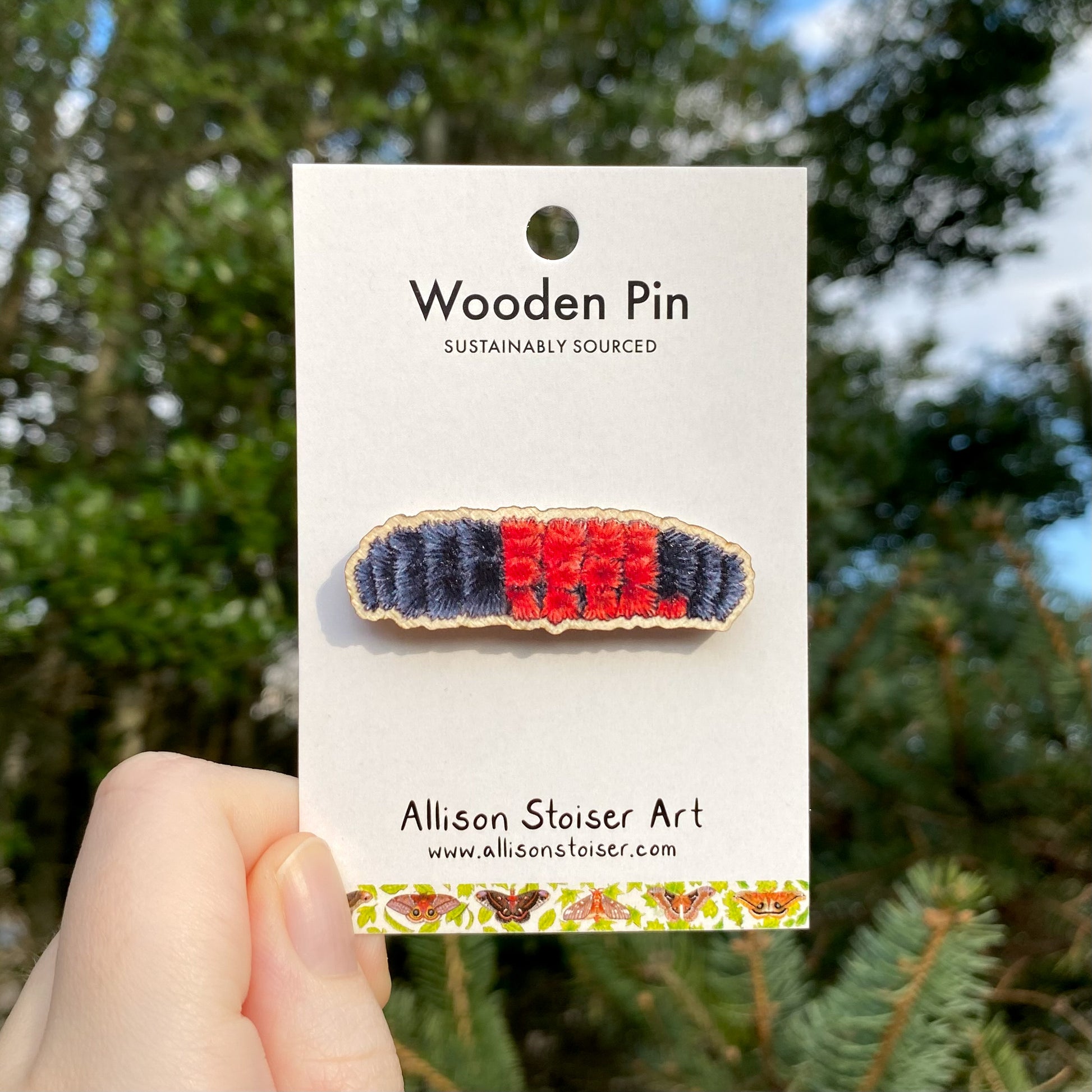 A wooden pin with a woolly bear caterpillar design, held by a hand, with a card that reads 'Wooden Pin Sustainably Sourced' from Allison Stoiser Art.