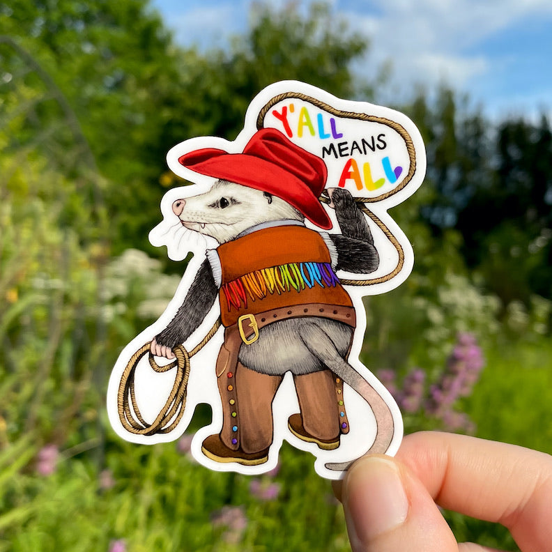 A hand holding a vinyl sticker of an opossum with a lasso dressed up in a rainbow cowboy outfit. In the lasso, it reads "Y'all means All" in rainbow text.