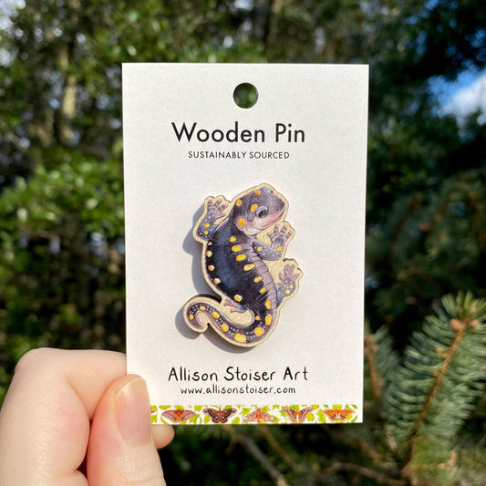 A hand holding a wooden pin with an illustration of a yellow-spotted salamander on it, labeled 'Wooden Pin Sustainably Sourced' by Allison Stoiser Art on the packaging.