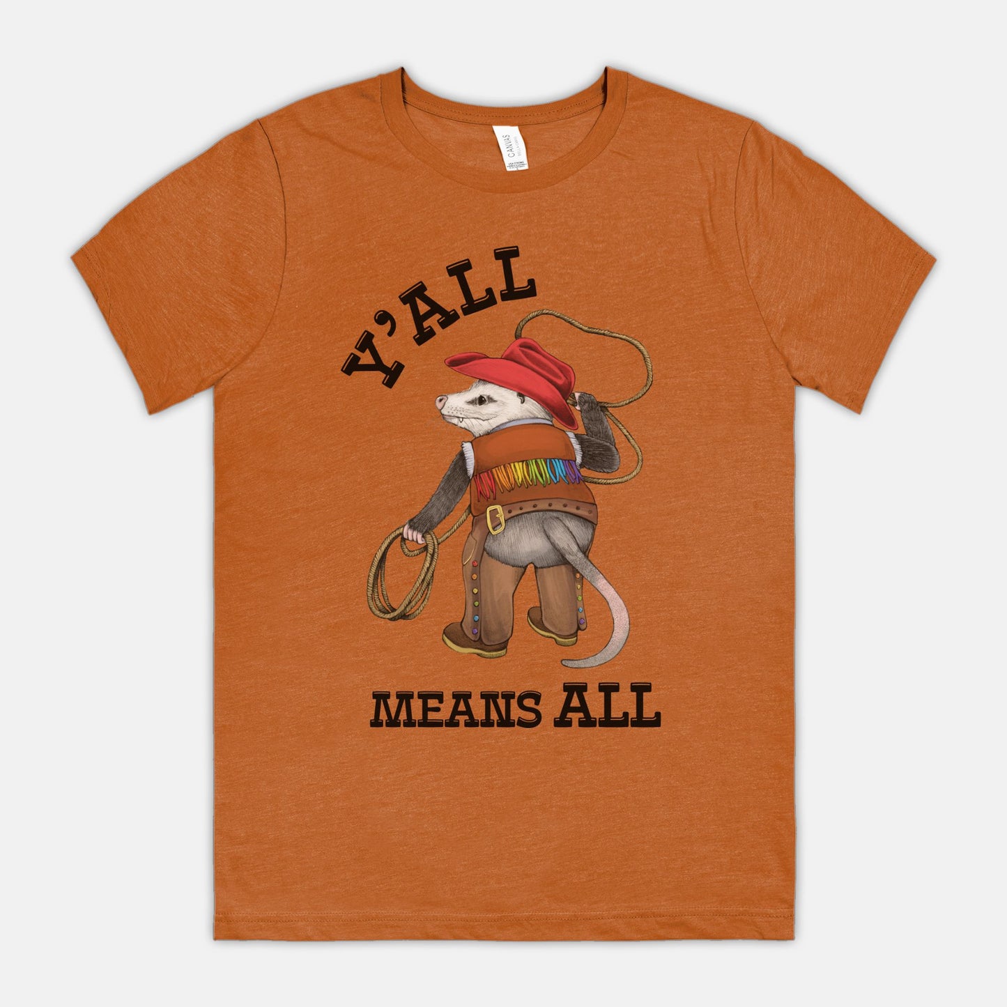 A dark orange-colored shirt featuring a cowboy opossum and text reading “Y’all Means All”