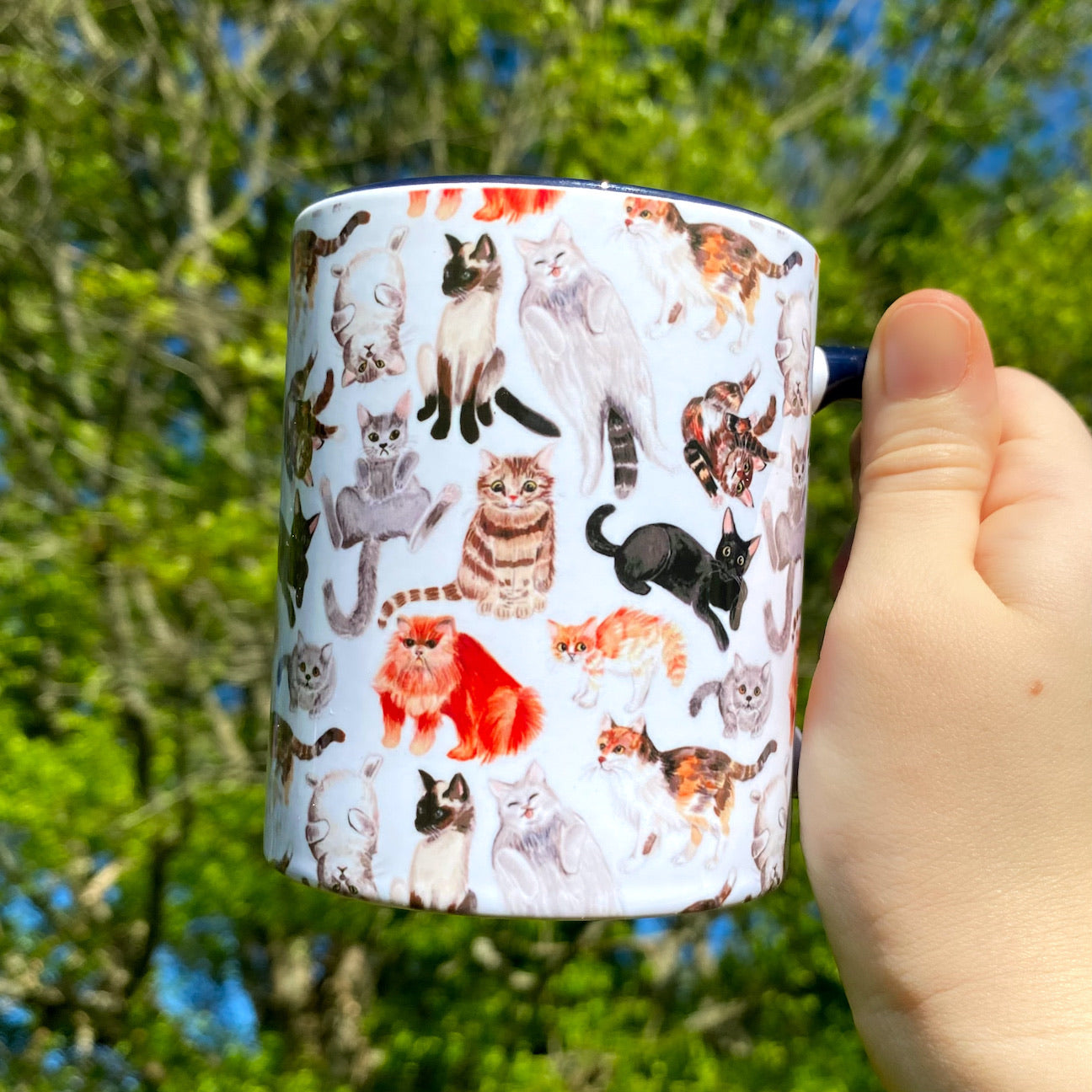 A hand holding a black handled mug with a watercolor cat pattern.