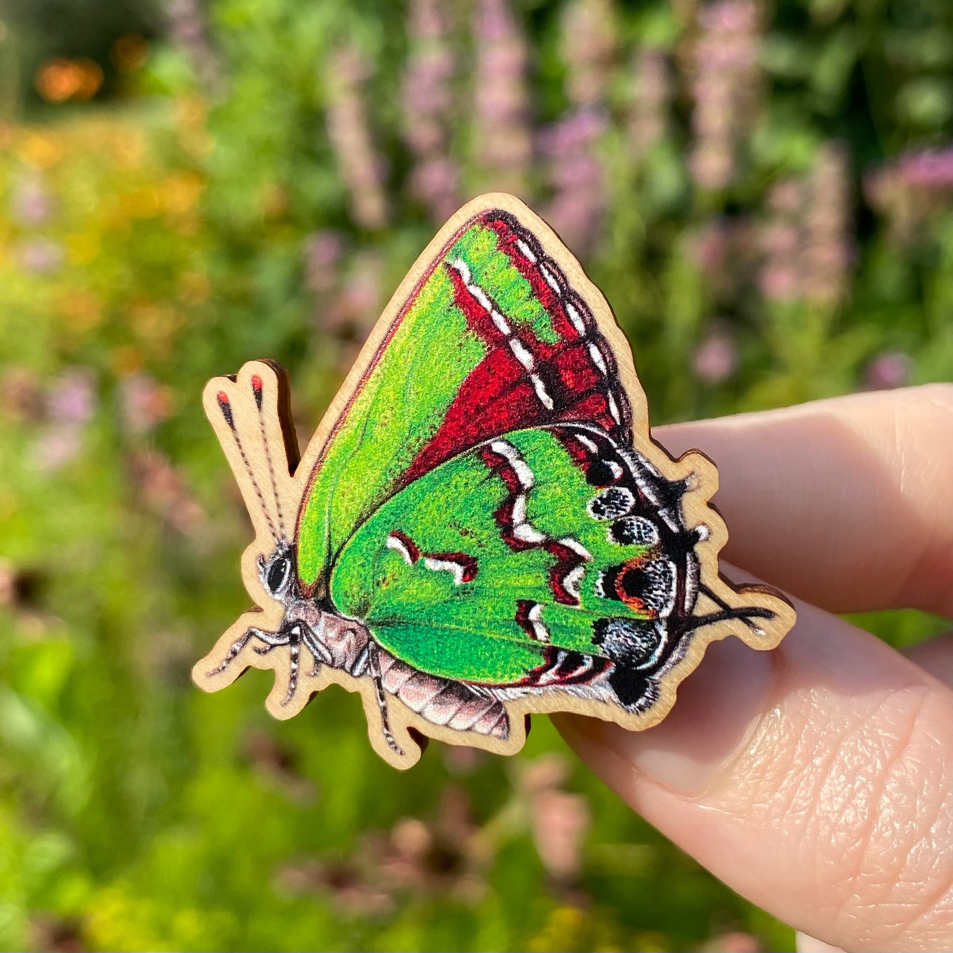 A hand holding a sustainably sourced wooden pin featuring an illustration of a juniper hairstreak