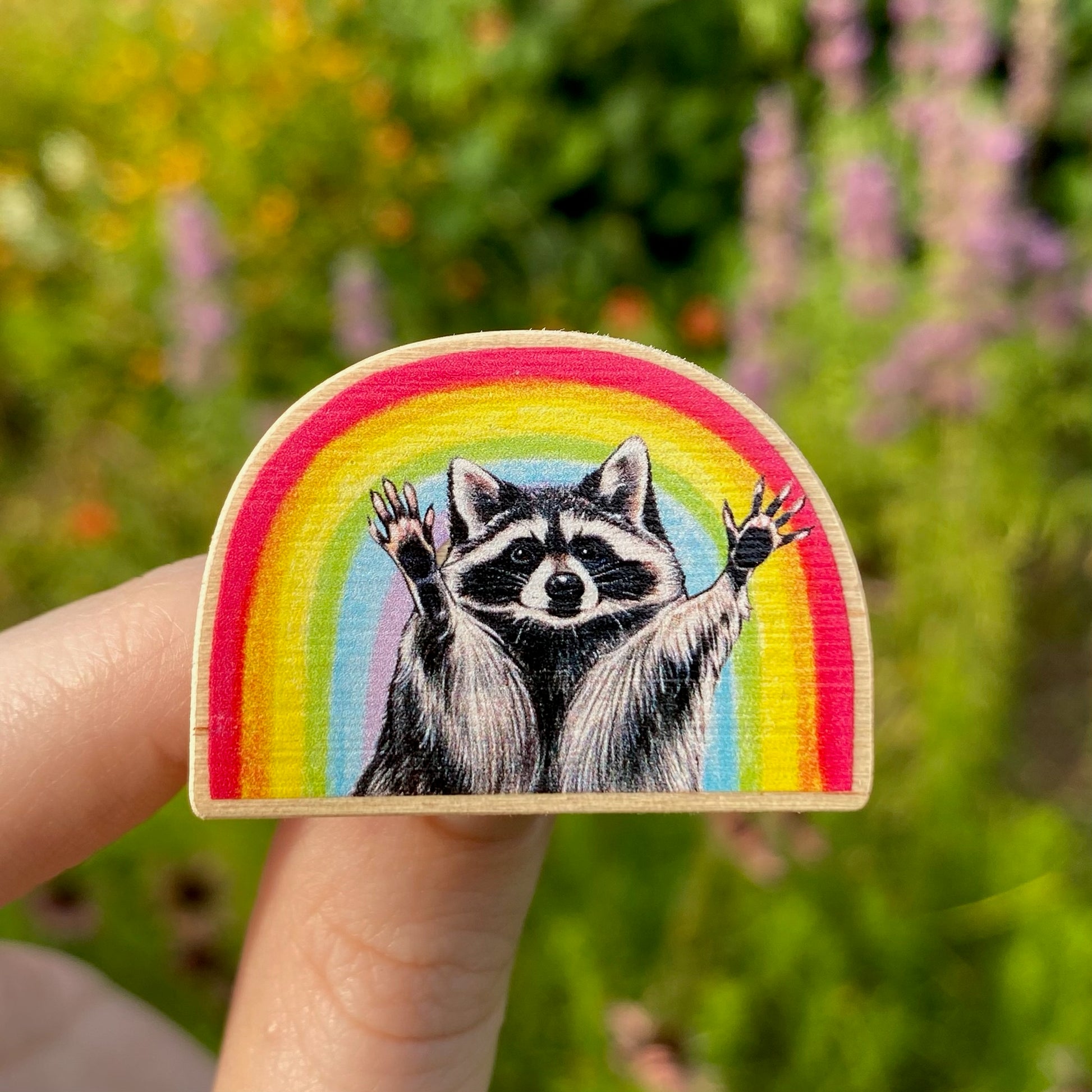 A hand holds a sustainably sourced wooden pin featuring an illustration of a raccoon with raised hands posed in front of a rainbow