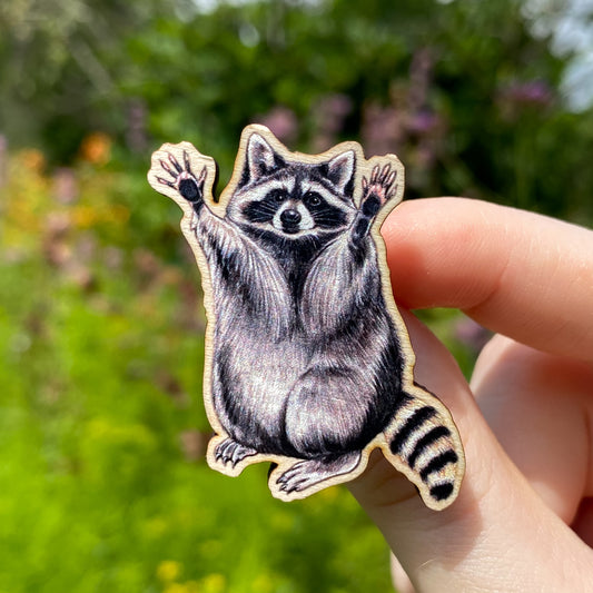 A hand holds a sustainably sourced wooden pin featuring an illustration of a raccoon