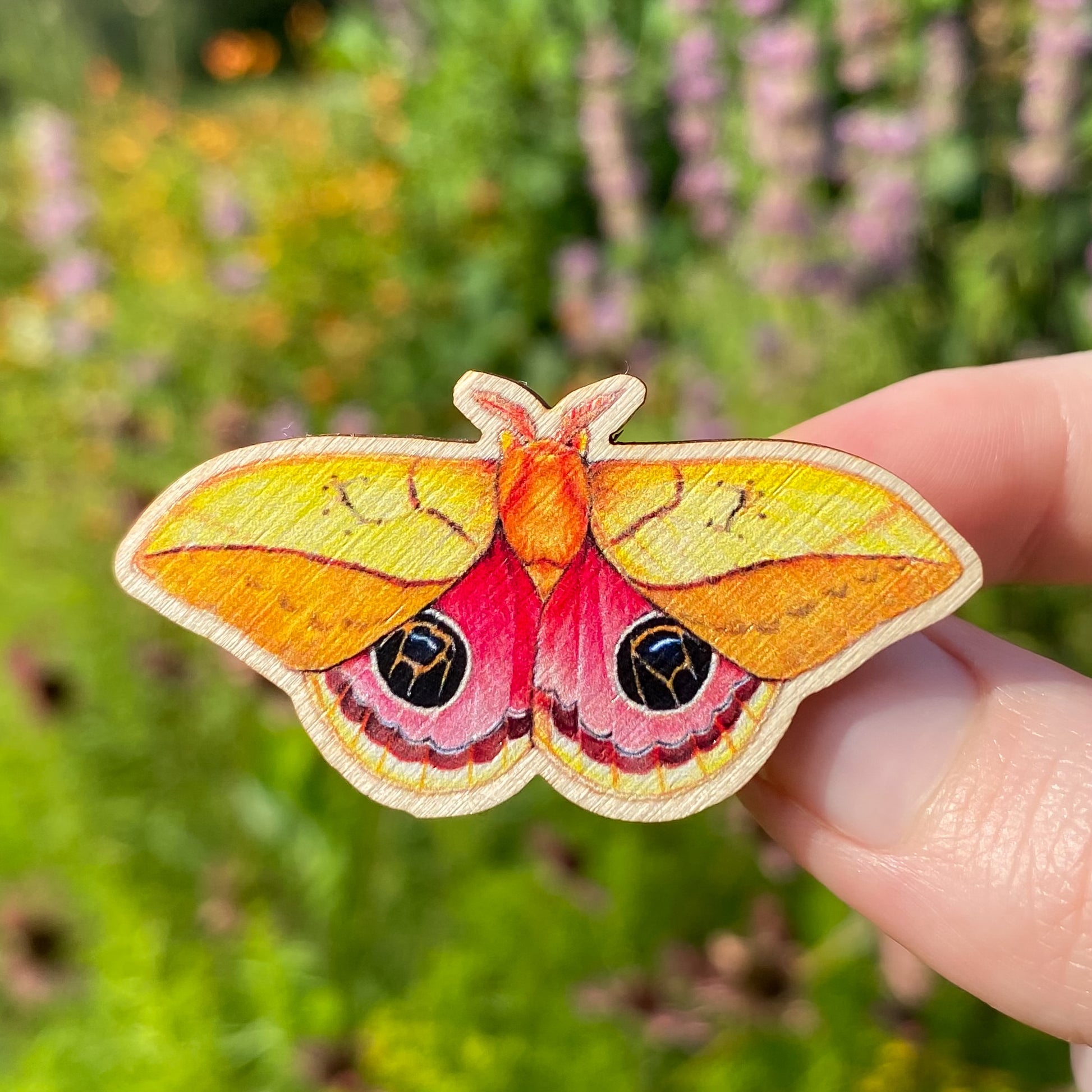 A hand holding a sustainably sourced wooden pin featuring an illustration of an Amazonian silkmoth