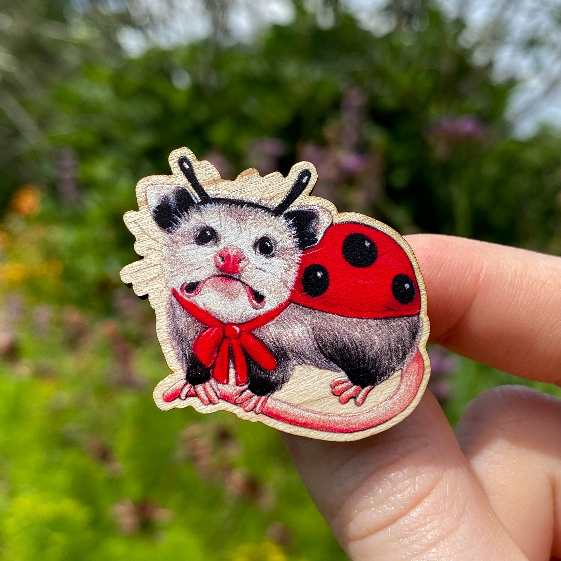 A sustainably sourced wooden pin featuring an opossum dressed as ladybug