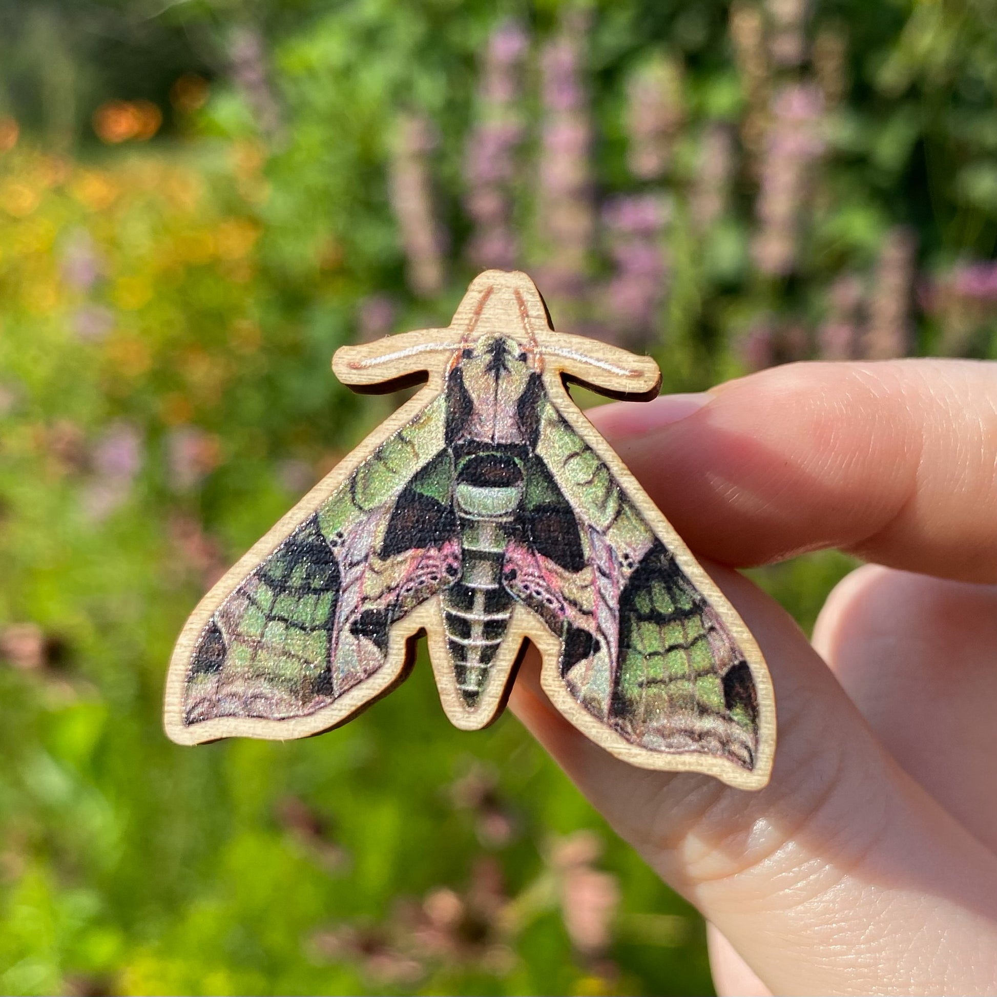 A hand holding a sustainably sourced wooden pin featuring an illustration of a pandorus sphinx moth