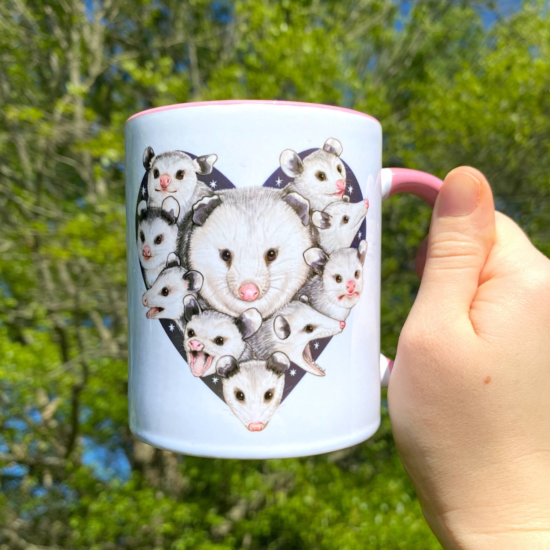 A hand holding a pink handled mug with a design of a mom opossum surrounded by her babies with a starry heart background.